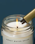 The gold candle lighter on a BLUE background that has the company "SWEET WATER DECOR" written on it lighting a white candle with"SWEET WATER DECOR" written on it 