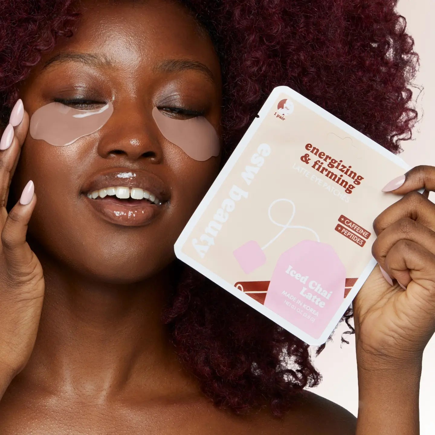 A woman with under-eye patches holds a package of ESW Beauty Energizing &amp; Firming Iced Chai Latte face masks. She has dark red curly hair, closed eyes, and is holding the product package in front of her face against a light beige background.
