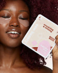 A woman with under-eye patches holds a package of ESW Beauty Energizing & Firming Iced Chai Latte face masks. She has dark red curly hair, closed eyes, and is holding the product package in front of her face against a light beige background.