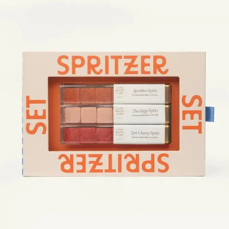 A boxed makeup spray set labeled SPRITZER SET, with a clear window showing three rectangular spray tubes in neutral and pink shades. The packaging is light beige with bold orange text, photographed on a clean white background.