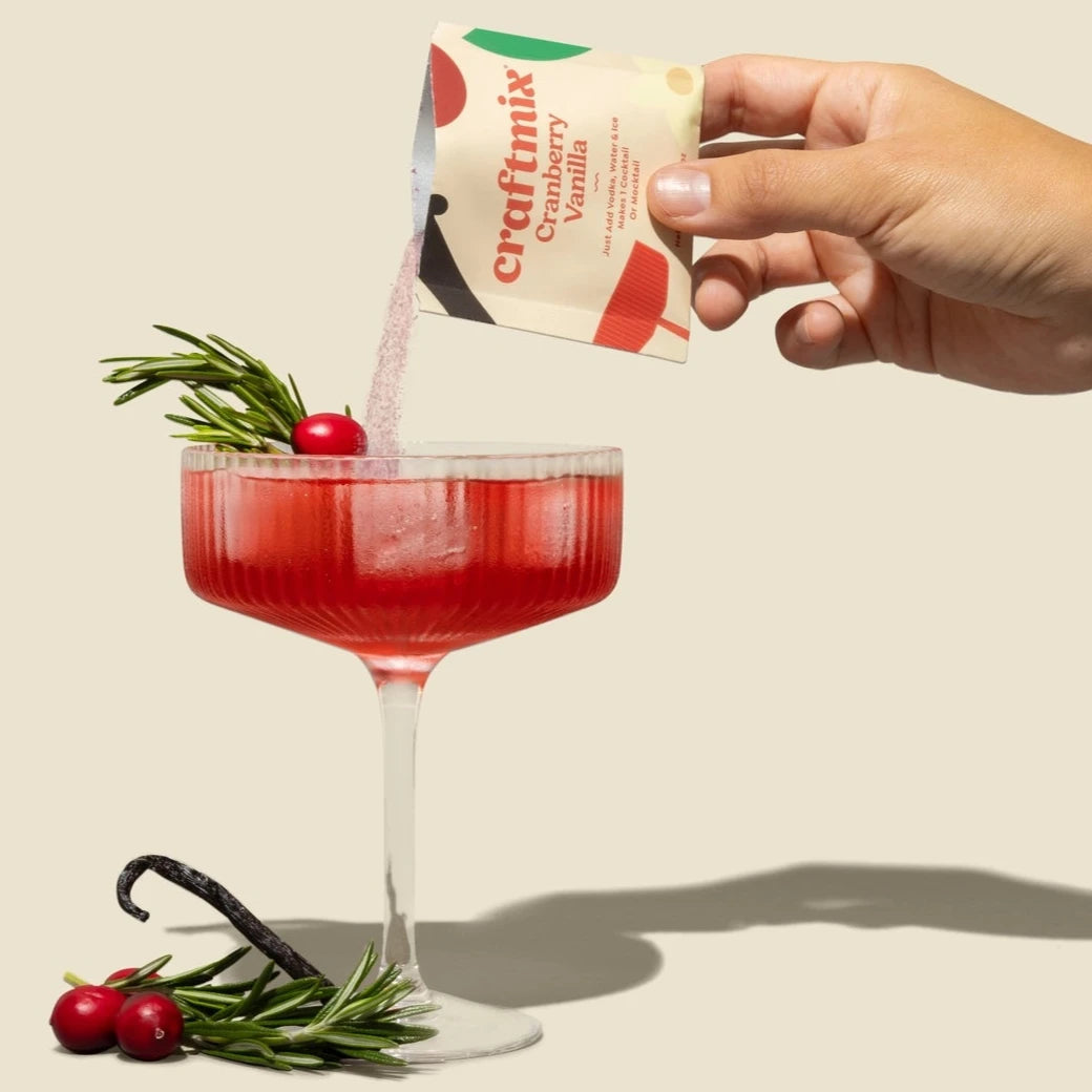 pouring drink mix into coupe filled with red liquid and garnished with rosemary and cranberries