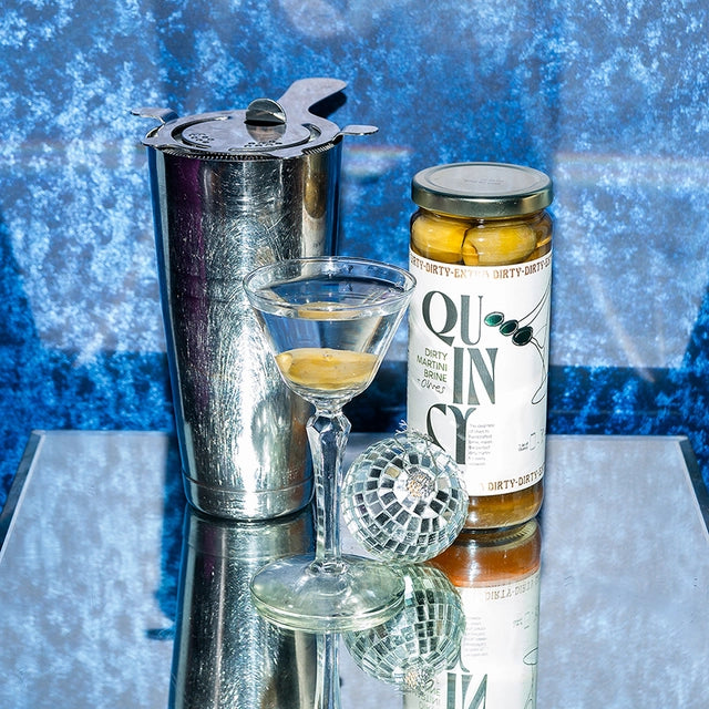 Cocktail shaker, glass with cocktail, and a bottle of Quin Sy on a reflective surface with a blue textured background.