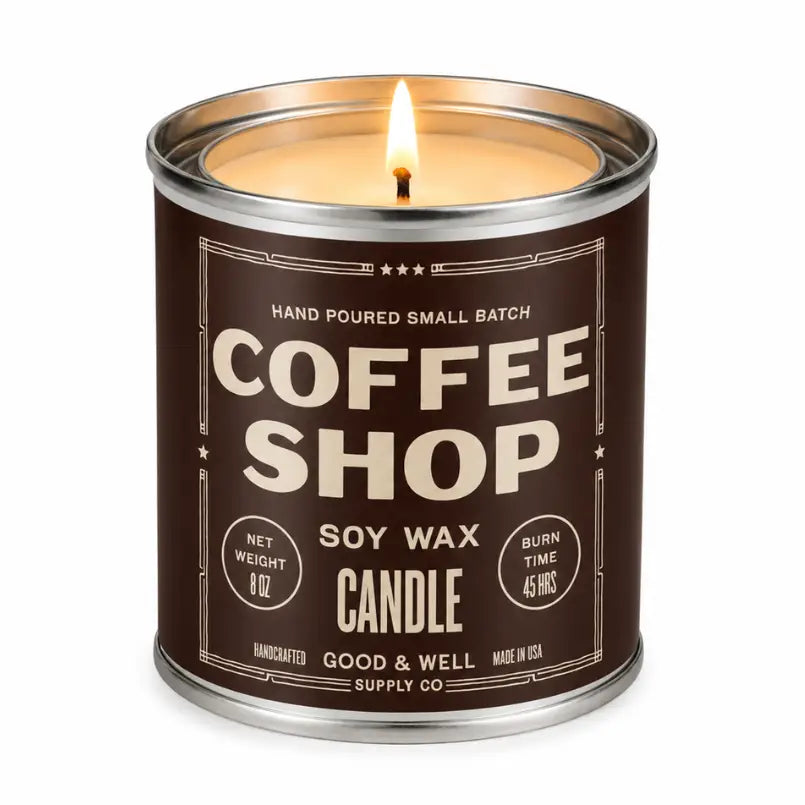 A lit soy wax candle in a cylindrical metal tin with a dark brown label reading 'COFFEE SHOP' and 'SOY WAX CANDLE' by Good &amp; Well Supply Co.