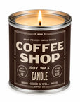 A lit soy wax candle in a cylindrical metal tin with a dark brown label reading 'COFFEE SHOP' and 'SOY WAX CANDLE' by Good & Well Supply Co.