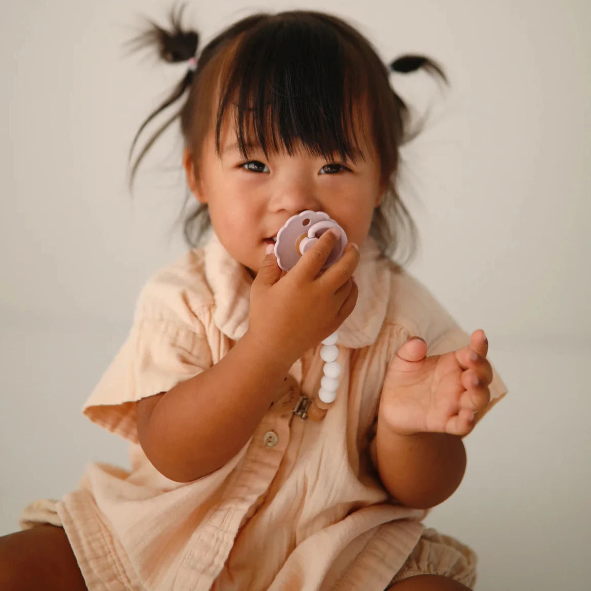 A young child with pigtails wearing a light beige outfit, holding and playing with a lilac-colored pacifier in hand, smiling softly towards the camera