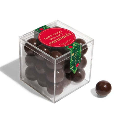 A clear acrylic cube box filled with dark chocolate sea salt caramels, with a red circular label and green ribbon, on a white background with one caramel placed outside the box.
