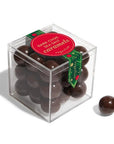 A clear acrylic cube box filled with dark chocolate sea salt caramels, with a red circular label and green ribbon, on a white background with one caramel placed outside the box.