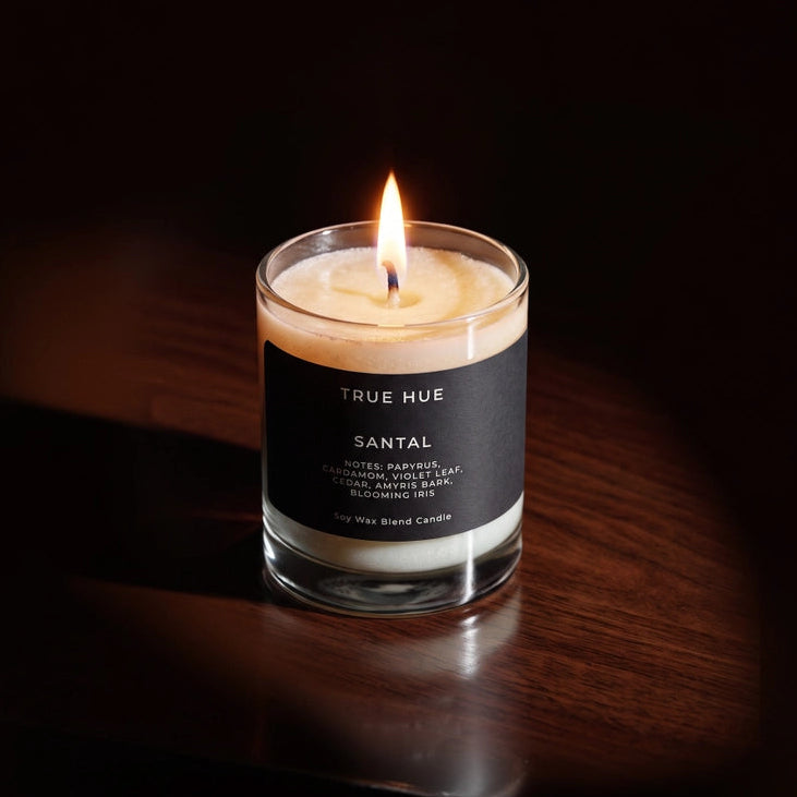 A lit candle in a clear glass jar with a black label reading TRUE HUE and SANTAL, placed on a dark wooden surface against a very dark background.
