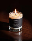 A lit candle in a clear glass jar with a black label reading TRUE HUE and SANTAL, placed on a dark wooden surface against a very dark background.