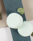 Two green bath bombs on a textured surface with leaves around
