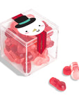 A clear acrylic cube container filled with red and pink gummy candies, with a snowman-themed paper label on top. Three loose gummies are placed in front on a clean white background.