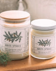 Two White Spruce candles on a wooden board with a neutral background