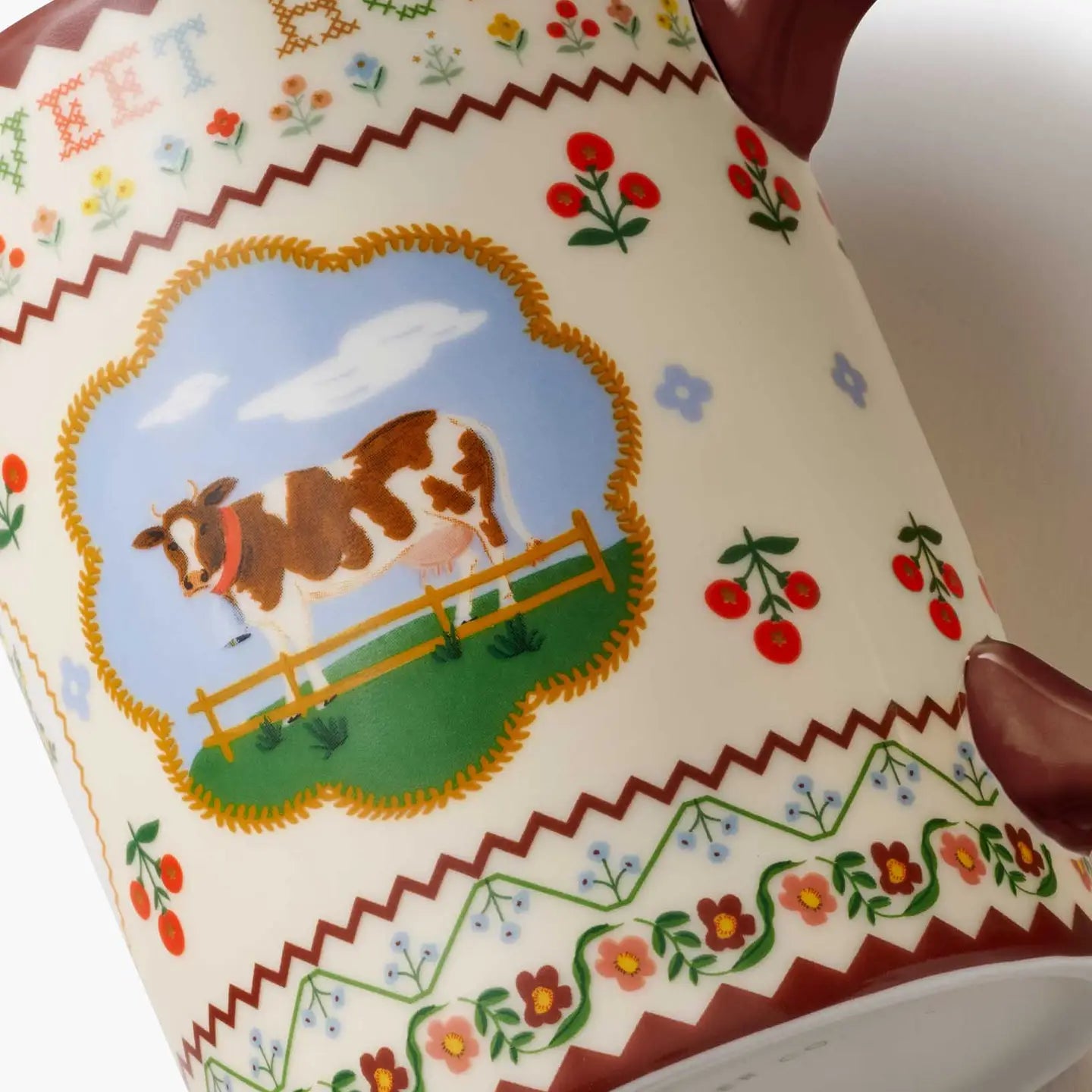 Close-up of a ceramic mug or cup with a cream-colored glaze, decorated with a colorful pastoral scene featuring a brown and white cow in a field, surrounded by floral and zigzag patterns.