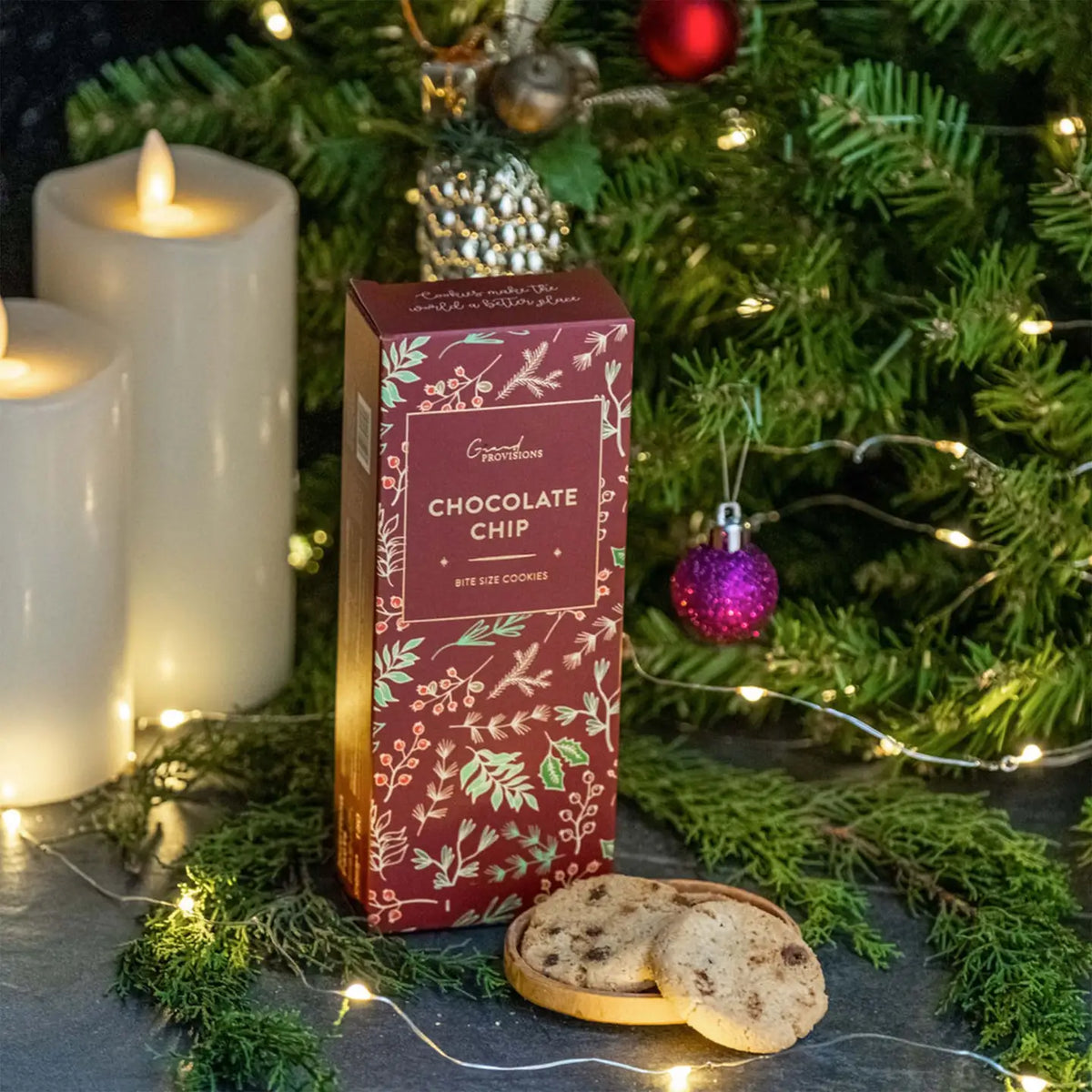 A box of Gifford's Chocolate Chip cookies stands on a dark surface in a festive holiday setting with lit candles, evergreen branches, string lights, ornaments, and two cookies in front.