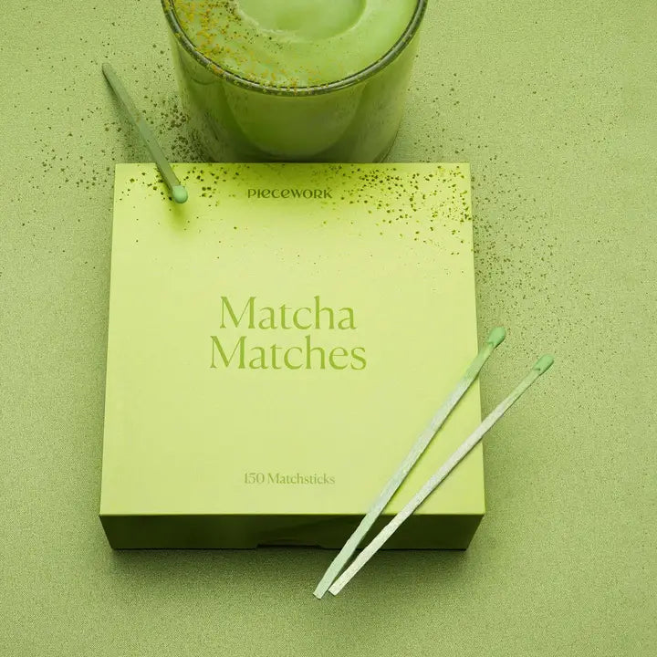 Box of 'Matcha Matches' with two matchsticks on a green surface, next to a glass of matcha.