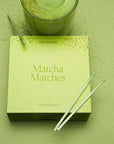 Box of 'Matcha Matches' with two matchsticks on a green surface, next to a glass of matcha.