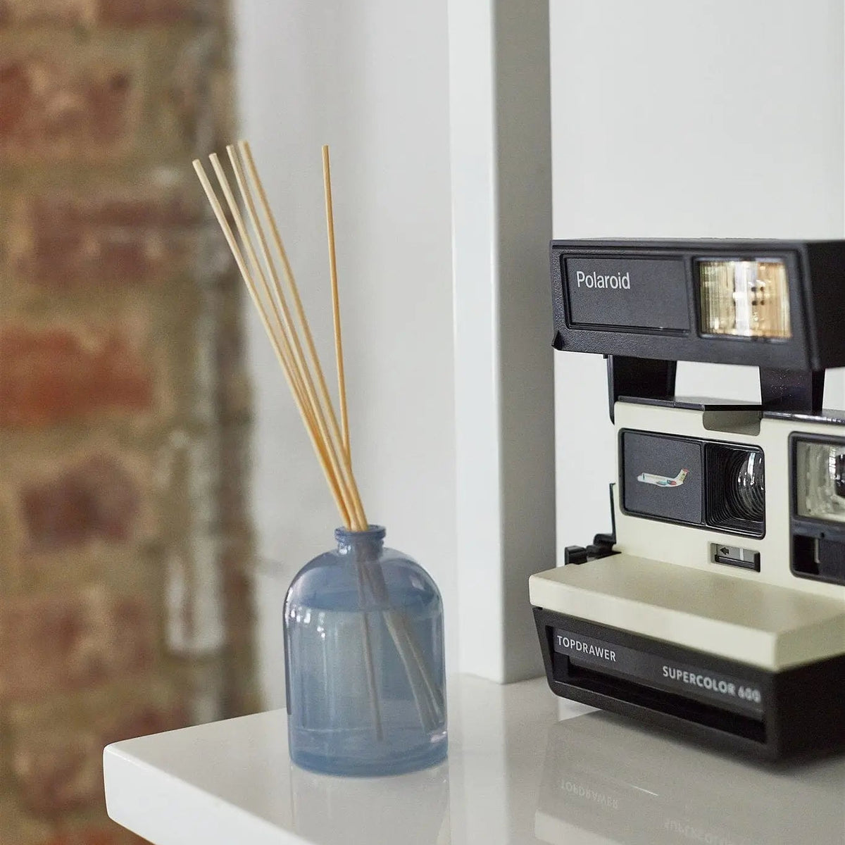 A blue glass reed diffuser bottle with natural reed sticks sits on a white shelf next to a vintage Polaroid camera. The background features a white wall and a blurred brick wall.