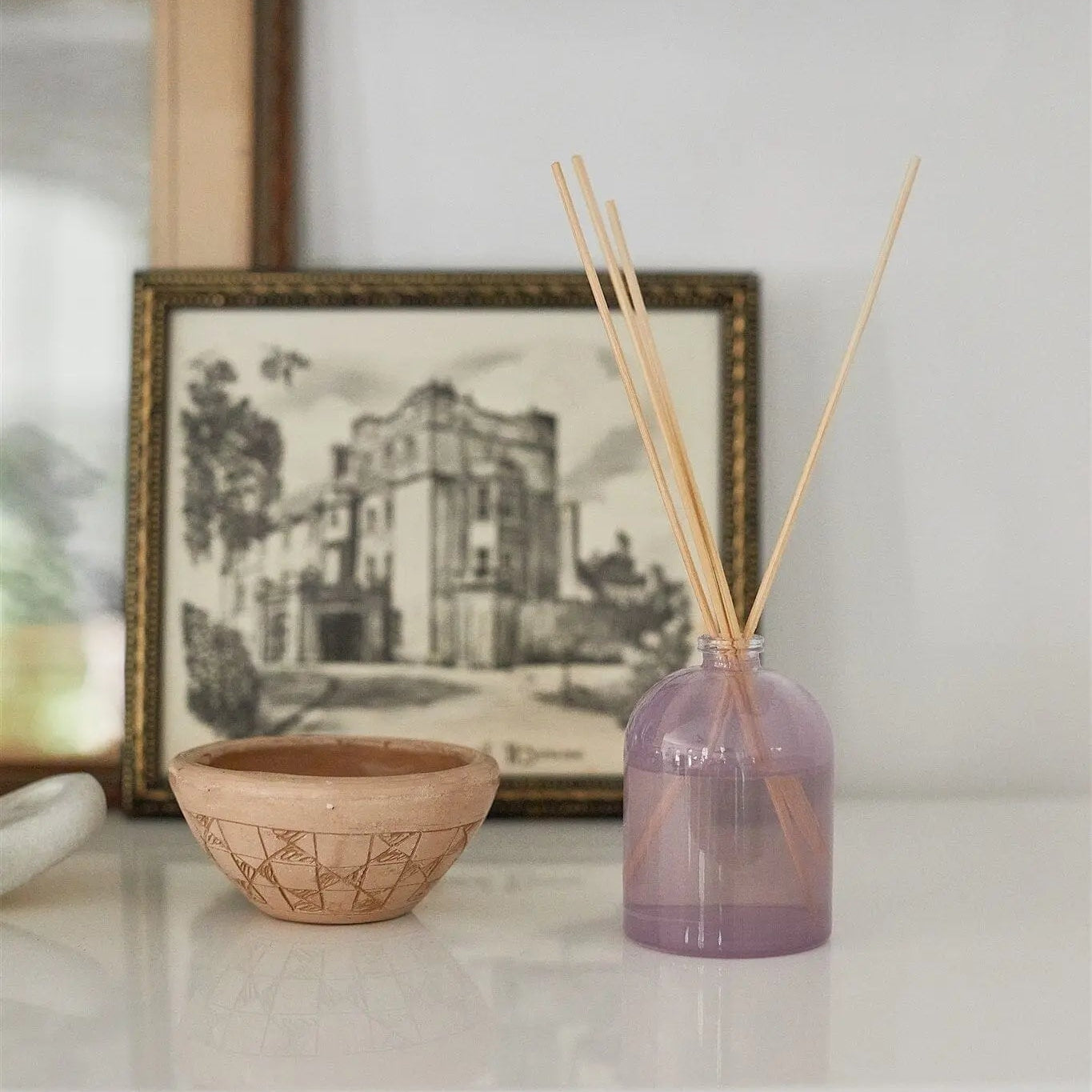 A lavender purple glass reed diffuser bottle with natural wood reeds on a white shelf, styled next to a small woven bowl and a framed print.
