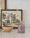 A lavender purple glass reed diffuser bottle with natural wood reeds on a white shelf, styled next to a small woven bowl and a framed print.