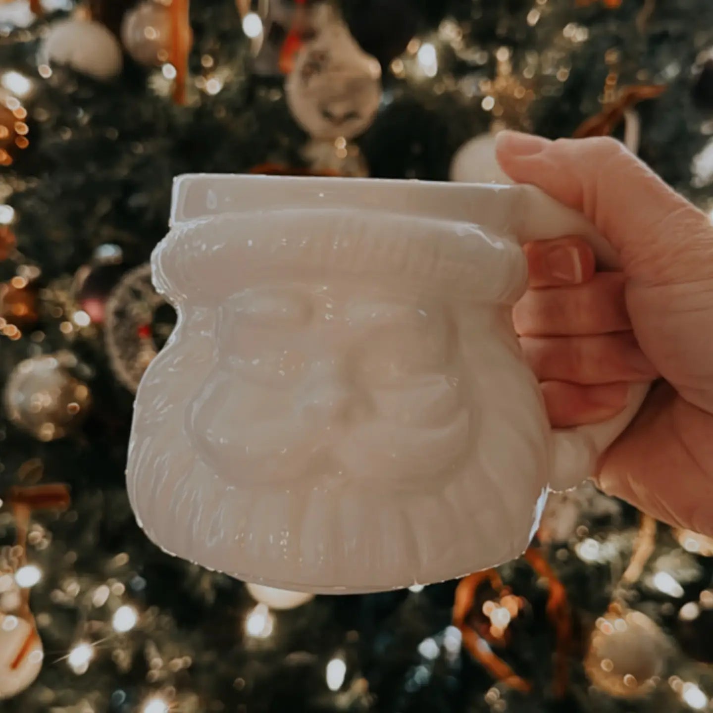 Hand holding a white ceramic mug with a Christmas tree in the background