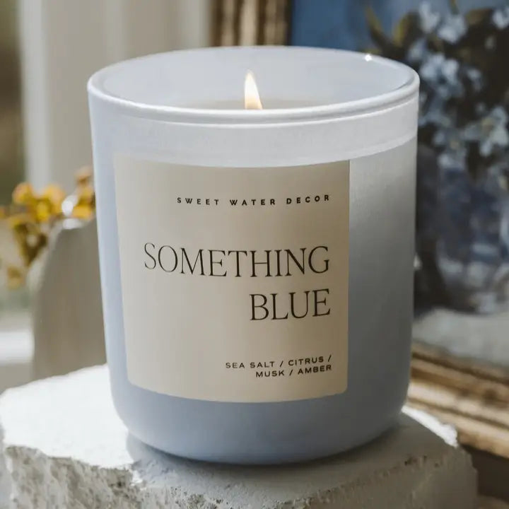 Candle labeled 'Something Blue' by Sweet Water Decor on a stone surface with a blurred background.