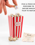 Red and white striped popcorn container with a piece of popcorn being picked, on a white background.