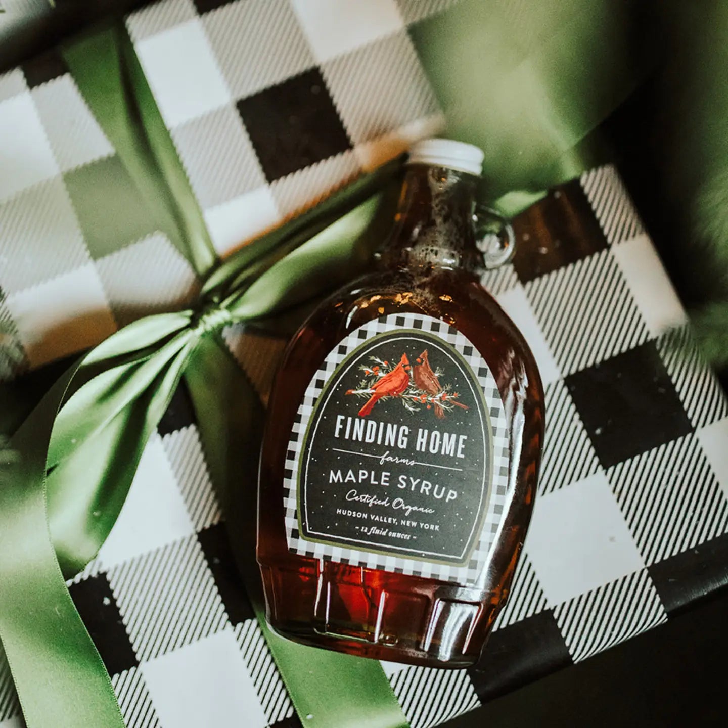 Bottle of Finding Home Maple Syrup on a checkered surface with green leaves.
