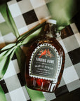 Bottle of Finding Home Maple Syrup on a checkered surface with green leaves.