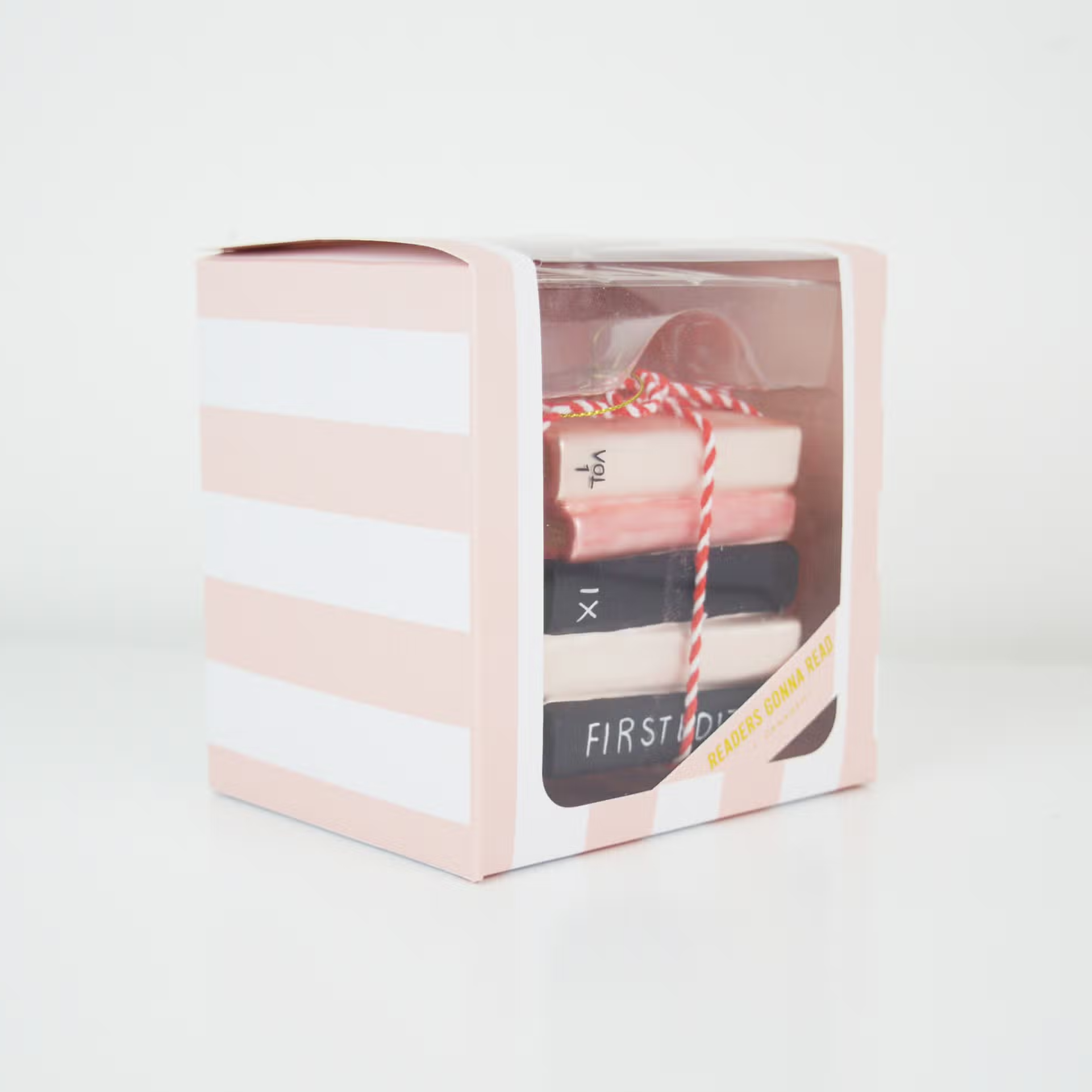 ornament shaped like a stack of book tied together with red and white twine. Book are pink and black with gold glitter showing the pages. Ornament is inside pink and white striped box