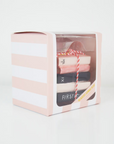 ornament shaped like a stack of book tied together with red and white twine. Book are pink and black with gold glitter showing the pages. Ornament is inside pink and white striped box