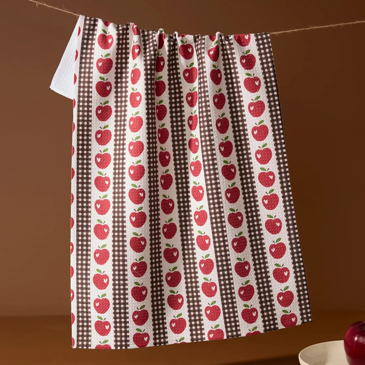 A kitchen curtain or hanging panel with a repeating red apple print on brown and white striped vertical bands, hanging from a rope against a warm brown background.