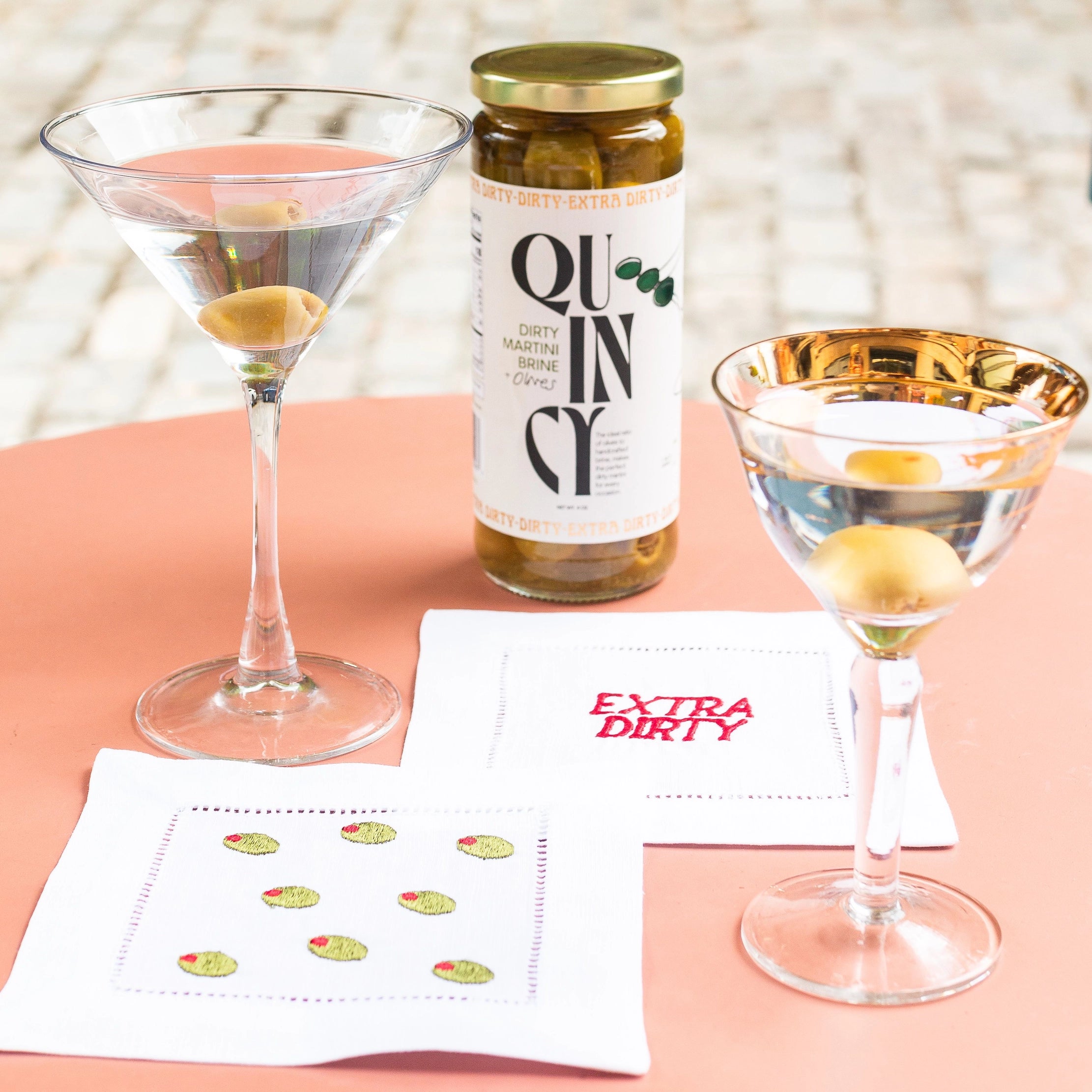 Two martini glasses with olives, a bottle labeled 'Quincy', and a napkin on a pink surface.