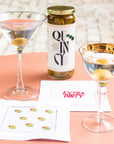 Two martini glasses with olives, a bottle labeled 'Quincy', and a napkin on a pink surface.
