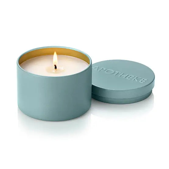 A teal blue metal candle tin with a lit wick and cream-colored wax, shown next to its matching lid which features the brand name 'APOTHEKE' embossed on a clean white background.