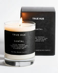 A lit TRUE HUE Santal soy wax blend candle in a clear glass jar with a black label, shown in front of its matching black product box on a clean white background.