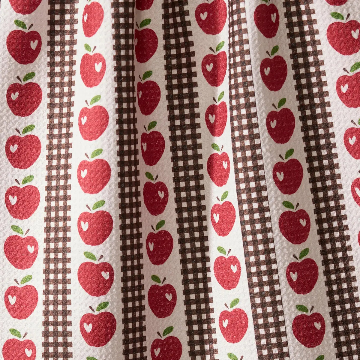 A close-up of a fabric pattern featuring vertical stripes with red apples, brown gingham, and a central brown-and-white checkered stripe on an off-white background.