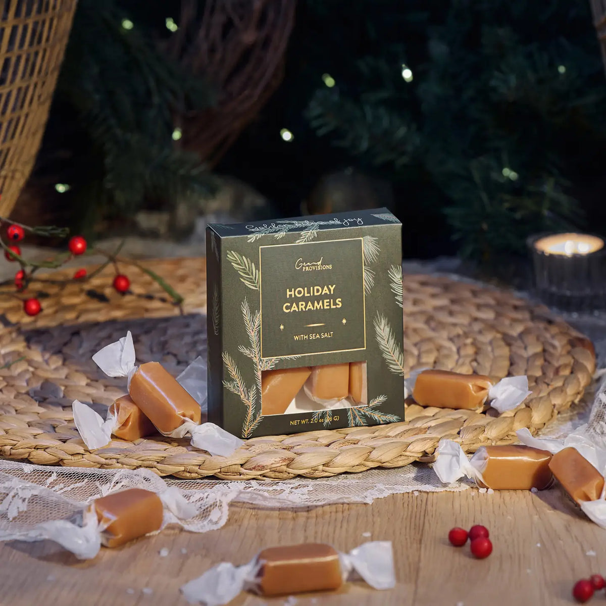 Box of Holiday Caramels with sea salt surrounded by wrapped caramels on a woven mat with a Christmas tree in the background.