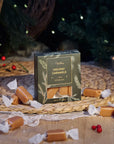 Box of Holiday Caramels with sea salt surrounded by wrapped caramels on a woven mat with a Christmas tree in the background.