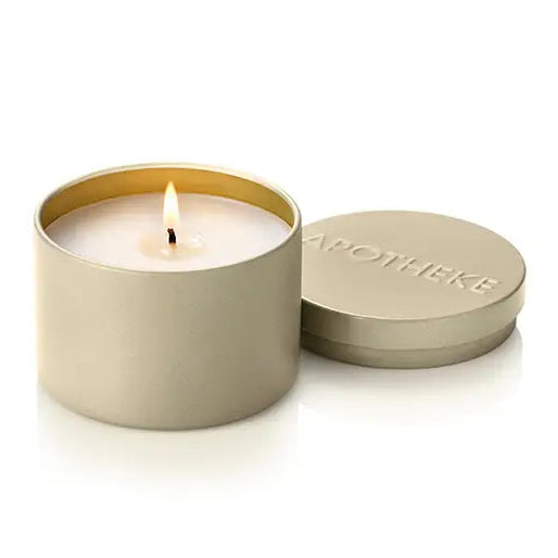 A lit cream-colored candle in a matte gold cylindrical tin with its lid resting beside it on a white background. The lid is embossed with the brand name 'APOTHEKE'.