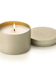 A lit cream-colored candle in a matte gold cylindrical tin with its lid resting beside it on a white background. The lid is embossed with the brand name 'APOTHEKE'.