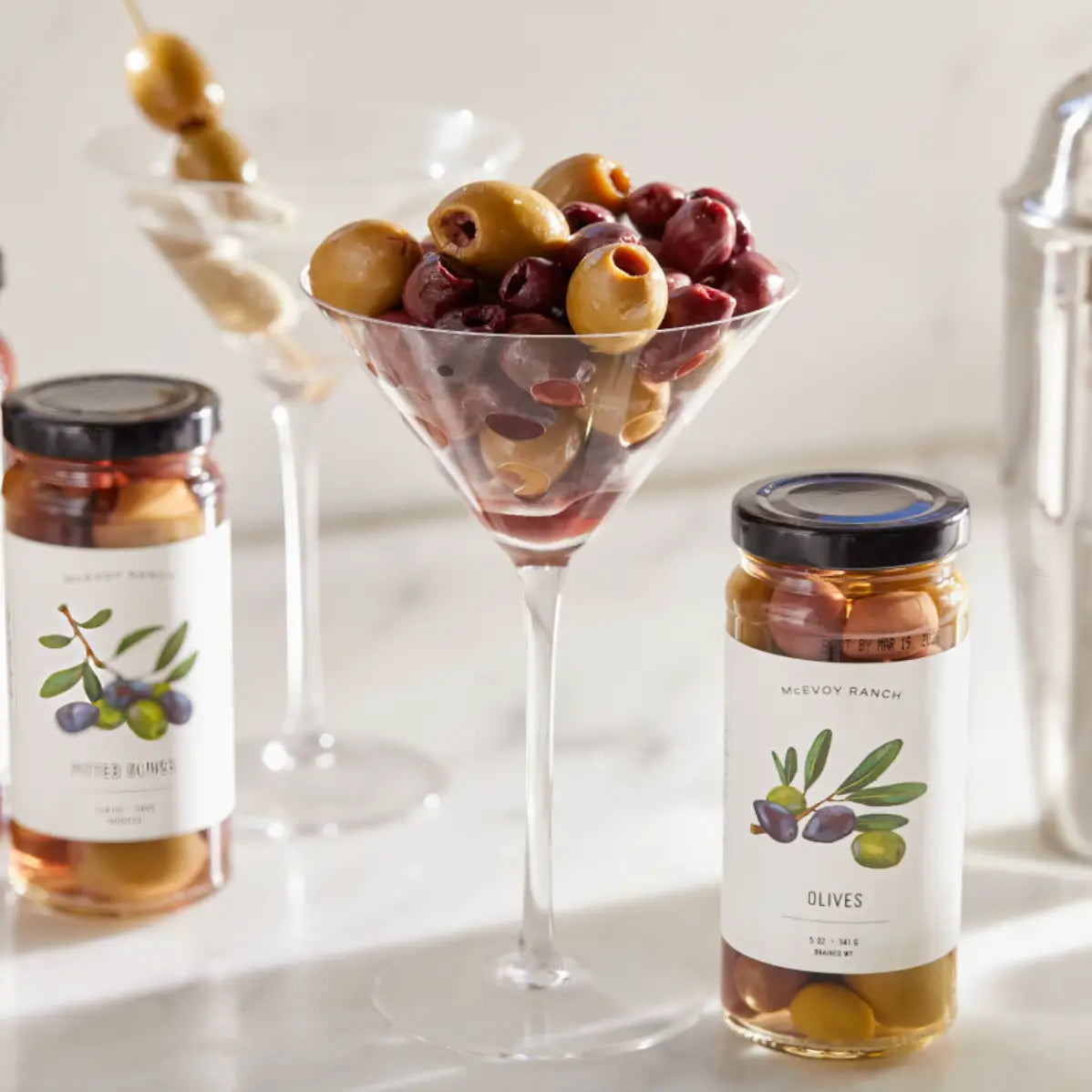 Two jars of olives with a martini glass filled with olives on a light background