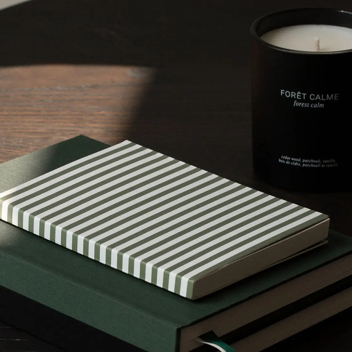 A striped notebook with white and dark green horizontal stripes sits on a dark green book, with a black candle labeled 'FORÉT CALME forest calm' in the background on a dark wooden surface.