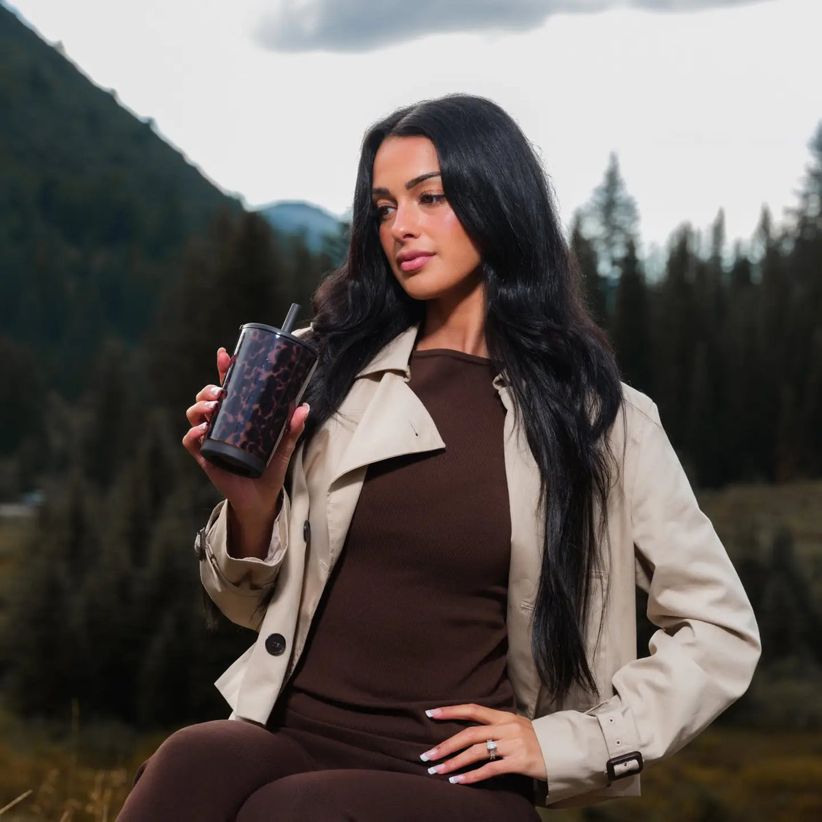 A woman with long dark hair sits outdoors in a mountain setting, wearing a beige trench coat over a brown ribbed top and brown leggings, while holding a dark patterned tumbler cup.