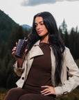 A woman with long dark hair sits outdoors in a mountain setting, wearing a beige trench coat over a brown ribbed top and brown leggings, while holding a dark patterned tumbler cup.