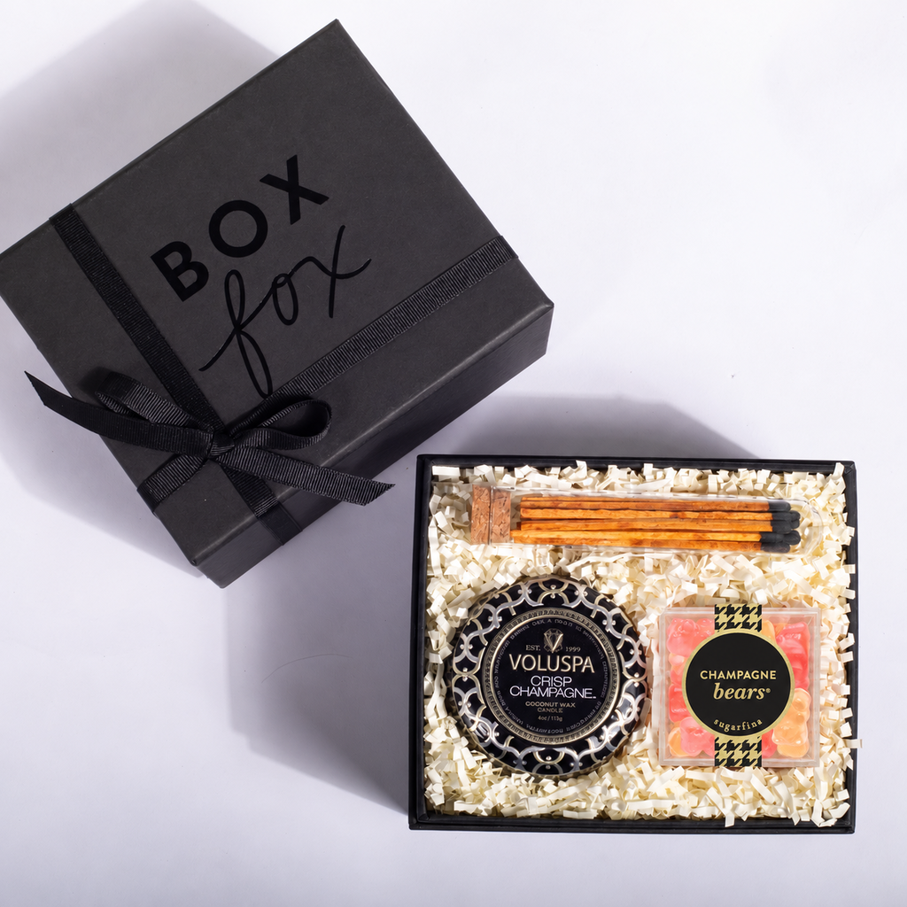 An open black gift box filled with shredded cream paper and spa items, including a black candle labeled Voluspa Champagne, a square soap bar with a champagne label, and two orange taper candles. A closed black gift box with 'BOX fox' printed on it sits beside the open box on a white background.