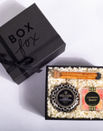 An open black gift box filled with shredded cream paper and spa items, including a black candle labeled Voluspa Champagne, a square soap bar with a champagne label, and two orange taper candles. A closed black gift box with 'BOX fox' printed on it sits beside the open box on a white background.