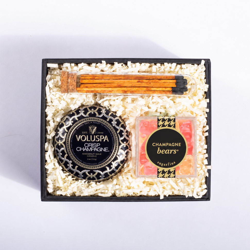A gift box set containing a Voluspa Crisp Champagne bottle cap, a pack of champagne buns, and a square box of 'Champagne bears' gummy bears, all arranged in a black tray filled with shredded white paper on a white background.