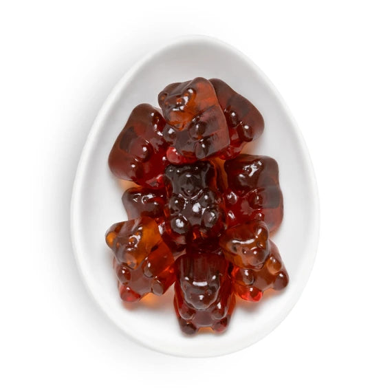 Gummy bear candies in a glossy amber-red translucent syrup, arranged on a white oval plate against a white background.