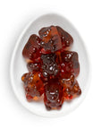 Gummy bear candies in a glossy amber-red translucent syrup, arranged on a white oval plate against a white background.
