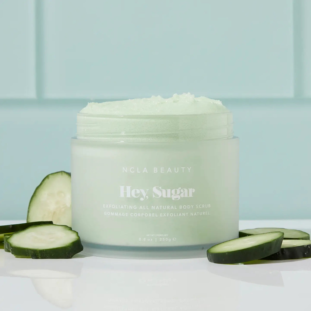 Jar of NCLA Beauty 'Hey Sugar' body scrub with cucumber slices on a light blue background